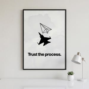 May include: Black framed print with a paper aeroplane above a fighter jet silhouette, and the text "Trust the process." The artwork is on a white surface, with a desk lamp and a small potted plant.