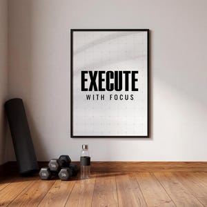 Puede incluir: Un póster enmarcado con las palabras "EXECUTE WITH FOCUS" en letras negras en negrita. Una esterilla de yoga negra, pesas y una botella de agua están en la esquina. El suelo es de madera.