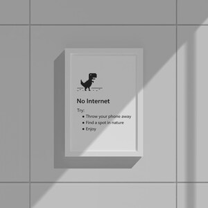 May include: A framed print with the text "No Internet" and a pixelated dinosaur graphic. The print suggests alternative activities: throw your phone away, find nature, and enjoy. The frame is white, and the background is a light gray wall.