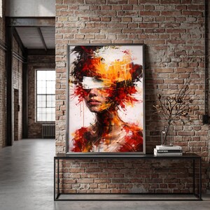 May include: Abstract portrait art print in a black frame. The artwork features a woman's face with a white band over the eyes and vibrant splashes of red, orange, and yellow paint. The print is displayed on a black metal table against a brick wall.
