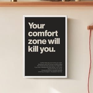 May include: A framed black and white poster with the text "Your comfort zone will kill you." The poster is in a white frame and hangs on a beige wall. Additional text is at the bottom of the poster.