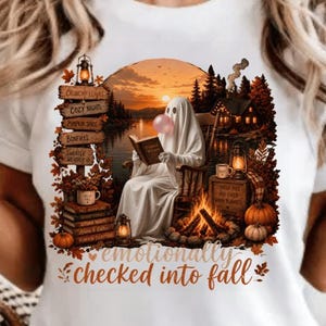 May include: A white t-shirt featuring a graphic print of a ghost sitting by a campfire, reading a book, and blowing a pink bubble. The design includes a wooden signpost with text, pumpkins, and the script text "emotionally checked into fall."