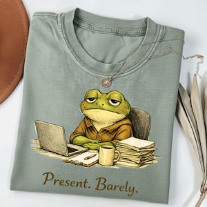 Könnte beinhalten: Salbeigrünes T-Shirt mit einer Cartoon-Fröschin an einem Schreibtisch mit Laptop, Kaffeetasse und Papieren. Die Fröschin hat einen müden Ausdruck. Der Text "Present. Barely." ist unten in brauner Schrift gedruckt.