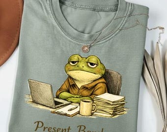 May include: Sage green t-shirt featuring a cartoon frog at a desk with a laptop, coffee mug, and paperwork. The frog has a weary expression. The text "Present. Barely." is printed in brown script at the bottom.
