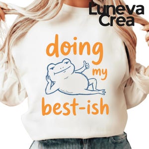 May include: White sweatshirt with the text "doing my best-ish" in orange and a cartoon frog illustration in blue. The frog is lounging with a thumbs-up gesture. The brand name "Luneva Crea" is printed in black at the top.