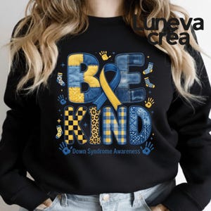 Puede incluir: Sudadera negra con las palabras "BE KIND" en letras de patchwork y una cinta azul y amarilla. El diseño incluye huellas de manos, calcetines y el texto "Down Syndrome Awareness". La sudadera tiene cuello redondo.
