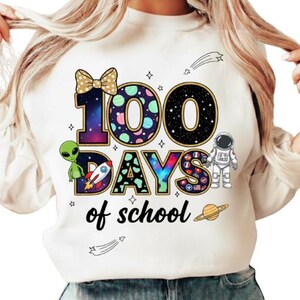 May include: White sweatshirt with a colorful graphic celebrating 100 days of school. The design includes the number "100" with a bow, an alien, a rocket, an astronaut, and the words "days of school". The graphic has a space theme with stars and planets.