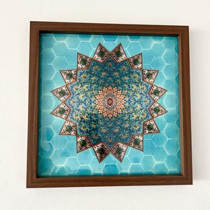 Può includere: Opera d'arte quadrata incorniciata con un design simmetrico dettagliato nei toni del turchese, oro e verde. Il motivo centrale è una stella, circondata da forme geometriche su uno sfondo di piastrelle esagonali turchesi. La cornice è in legno marrone scuro.