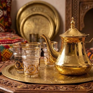 May include: A golden tea set with a teapot, glasses, and a tray. The teapot and tray have intricate designs. The glasses have gold rims and floral patterns. The set is on a carved wooden table, suggesting a cultural theme.