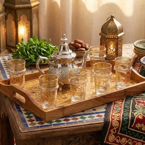 May include: A wooden serving tray displays a silver teapot, eight patterned glasses, and dates. The tray is on a table with a mosaic top, with lanterns and bowls, creating a warm, inviting atmosphere. The scene evokes a sense of hospitality.