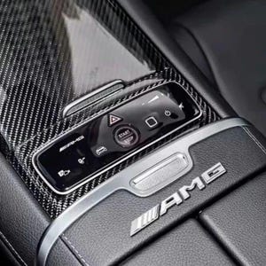 Puede incluir: Primer plano del interior de un coche con un panel de fibra de carbono, un panel de control y el botón "START STOP". El logotipo plateado "AMG" es visible en el reposabrazos de cuero negro.
