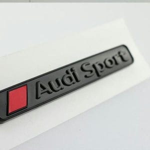 Könnte beinhalten: Schwarzes Audi Sport Emblem mit rotem Diamantakzent. Das rechteckige Abzeichen hat erhabene Schriftzüge und ist wahrscheinlich für ein Fahrzeug bestimmt. Das Emblem befindet sich auf weißem Hintergrund.
