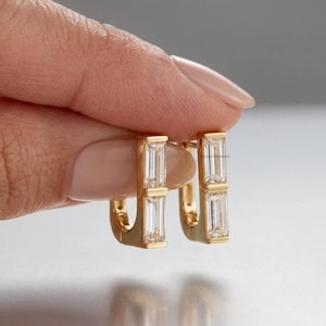 Puede incluir: Un par de pendientes de aro dorados con dos piedras transparentes de corte baguette apiladas verticalmente en cada pieza. Los pendientes tienen un acabado en oro pulido y un mecanismo de cierre de bisagra, con un diseño geométrico limpio.