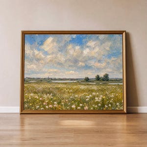 Op de afbeelding: Een ingelijst landschapsschilderij met een goudkleurige lijst. Het kunstwerk toont een veld met wilde bloemen onder een bewolkte blauwe lucht. Een waterpartij en een verre stad zijn zichtbaar op de achtergrond. Het schilderij heeft een vintage esthetiek.
