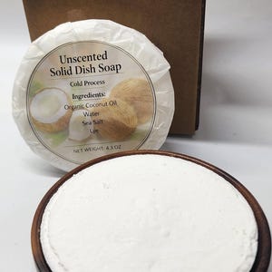 May include: A round white solid dish soap bar in a dark wood bowl, accompanied by a wrapped soap bar labeled Unscented Solid Dish Soap with coconut imagery and a net weight of 4.3 oz, plus a rectangular dual-textured brown and white sponge.