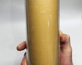 May include: A tall, cylindrical candle in a clear glass holder. The candle is a warm, golden color with a textured surface. The wick is visible at the top. The candle is held by a person's hand.
