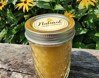 May include: A yellow candle in a clear glass jar with a silver lid. The jar has a diamond pattern. The label reads "Natural Light Candles". The background features yellow flowers and green foliage.