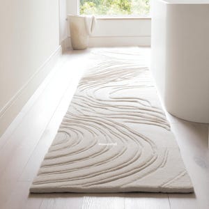 Op de afbeelding: Een lange, crèmekleurige vloerkleed met een getextureerd, wervelend patroon. Het rechthoekige kleed ligt op een lichtgekleurde houten vloer. Het ontwerp lijkt op waterstromen. Het kleed bevindt zich in een badkamer.
