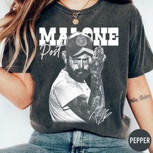 May include: Dark gray t-shirt featuring a black and white image of Post Malone. The word "MALONE" is printed in large white block letters above the image, with "Post" in a cursive font. The artist's signature is at the bottom.