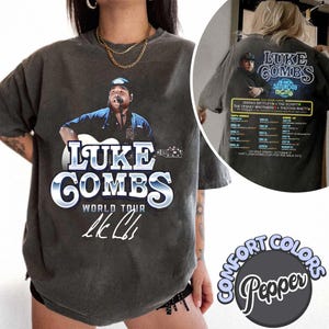 May include: Pepper-colored Comfort Colors t-shirt featuring a graphic of Luke Combs playing a guitar. The front design includes "LUKE COMBS WORLD TOUR" text and his signature. The back displays tour dates and artist names.