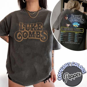 May include: Dark grey Comfort Colors t-shirt featuring the text "LUKE COMBS" in a brown, vintage-style font. The back of the shirt has a concert graphic with tour dates. The shirt is a Pepper color.