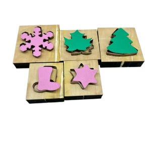 Könnte beinhalten: Eine Sammlung von Holzstanzformen in verschiedenen Formen, darunter eine rosa Schneeflocke, ein grünes Blatt, ein grüner Weihnachtsbaum, ein rosa Stiefel und ein rosa Stern. Die Stanzformen sind auf weißem Hintergrund angeordnet.