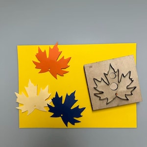 May include: A yellow rectangular card with three paper maple leaves in orange, cream, and navy blue. A wooden die-cut maple leaf with the number 100 is on the right side of the card. The background is gray.
