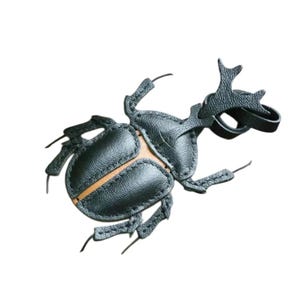 Peut inclure: Un ornement en forme de scarabée rhinocéros en cuir noir avec une surface texturée, des coutures visibles et une bande centrale beige. Le scarabée possède une corne proéminente et une petite boucle en cuir noir fixée sur le dessus pour l'accrocher.