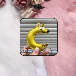 May include: A yellow leather keychain shaped like the letter C, designed to resemble a cartoon dog with white eyes, a black nose, and stitched edges. The item features a gold-toned metal grommet at the bottom with a thin cord attached for hanging.