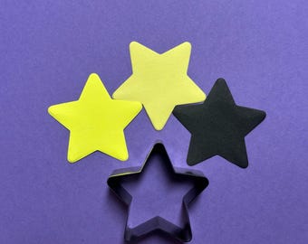 May include: A purple background with four star-shaped cutouts in yellow and black, along with a metal star-shaped cookie cutter. The stars are arranged in a visually appealing composition, suggesting a crafting or baking theme.