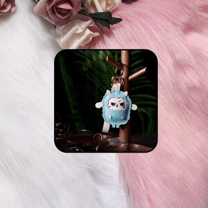 May include: A light blue leather keychain featuring a cartoon owl character with a grumpy expression, white scalloped trim, and two small pink dots. The owl is attached to a gold-toned metal ring and a light-colored strap, hanging from a wooden stand.