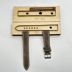 Może przedstawiać: Drewniana matryca do paska do zegarka z dwoma brązowymi paskami do zegarka zamszowego. Matryca jest oznaczona jako "18-16" i "8-16". Paski do zegarka są brązowe z białymi przeszyciami i srebrną klamrą.