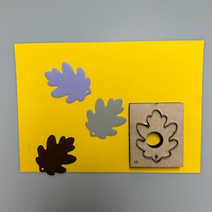 Peut inclure: Une carte jaune avec trois découpes de feuilles marron, grise et lavande, et une matrice en bois en forme de feuille. La matrice a un contour métallique et le chiffre 08.