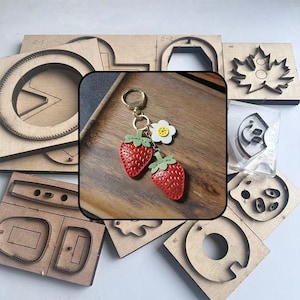 Könnte beinhalten: Ein roter Erdbeer-Schlüsselanhänger mit grünen Blättern und einem weißen Blumenanhänger, befestigt an einem goldenen Ring. Verschiedene hölzerne Stanzformen sind um den Schlüsselanhänger angeordnet.