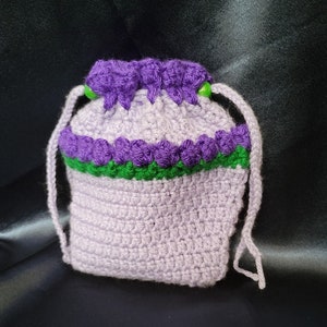 May include: A purple crocheted drawstring bag with green trim and green beads. The bag is made with a textured stitch and has a simple design.