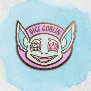 May include: A round enamel pin featuring a cartoon goblin face with the text "DICE GOBLIN". The goblin has light blue skin, large ears, and eyes shaped like polyhedral dice. The pin has a gold border and a pink and purple background.