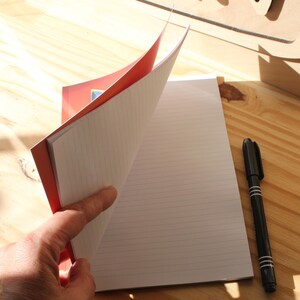 May include: A red and white lined notebook with a black pen on a wooden surface. The notebook is open, revealing the lined pages. The cover is red with a design. The pen is black with white stripes.
