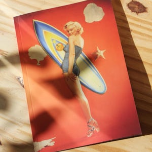May include: A notebook with a retro illustration of a woman in a swimsuit with a surfboard on a red background. The surfboard is blue and yellow, and the woman is blonde. The notebook has a glossy finish.