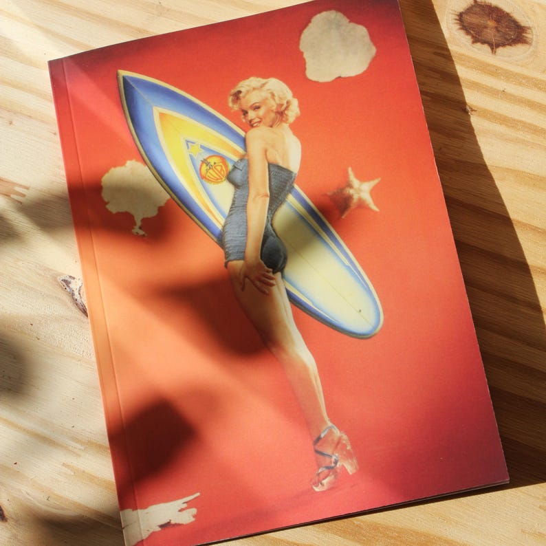 May include: A notebook with a retro illustration of a woman in a swimsuit with a surfboard on a red background. The surfboard is blue and yellow, and the woman is blonde. The notebook has a glossy finish.