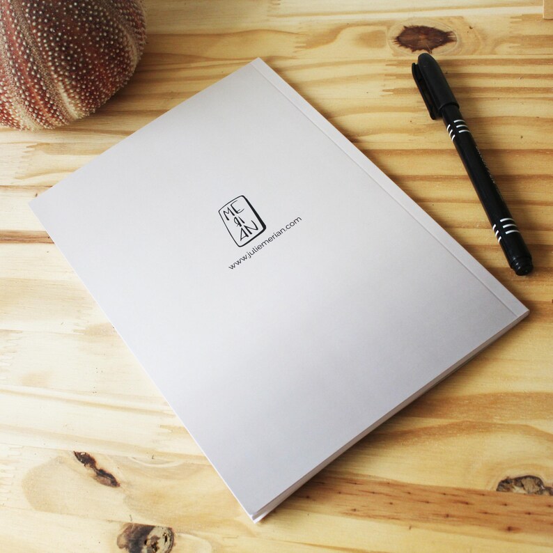 May include: A closed, light grey notebook with the text "MC AN" and "www.juliemerian.com" printed on the cover. A black pen with silver stripes lies next to the notebook on a wooden surface. A sea urchin is in the upper left corner.