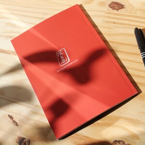May include: A red notebook with the text "MC AN" inside a square and the website "www.juliemenan.com". A black pen is visible on a wooden surface.
