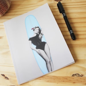 May include: A notebook with a black and white image of a woman in a black swimsuit leaning on a light blue surfboard. A black pen is placed on the wooden surface beside the notebook.