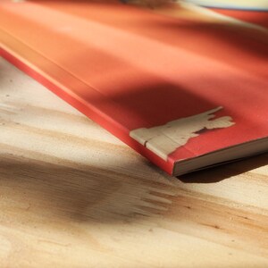 May include: A red book with a gradient cover, on a wooden surface. The book's corner shows exposed cardboard. The cover's colour transitions from dark red to light orange. The book is likely a paperback.