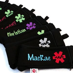 May include: A stack of black zippered pouches with a red, green, purple, and gray medical symbol and personalized names. The pouches are made by 'Left Wrist Wear'.