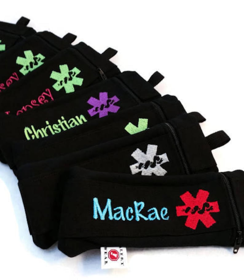 May include: A stack of black zippered pouches with a red, green, purple, and gray medical symbol and personalized names. The pouches are made by 'Left Wrist Wear'.