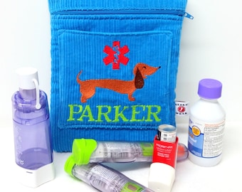 Op de afbeelding: Een blauwe corduroy ritszak met een rood medisch symbool en een teckel geborduurd op de voorkant. De tas heeft een groene zak en de naam "PARKER" is geborduurd onder de teckel. De tas ligt op een wit oppervlak met een inhalator, een fles vloeibaar medicijn en een waterfles.