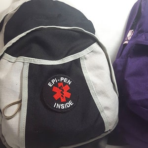 Puede incluir: Una mochila negra y gris con un parche blanco que dice "EPI-PEN INSIDE" con un símbolo médico rojo.