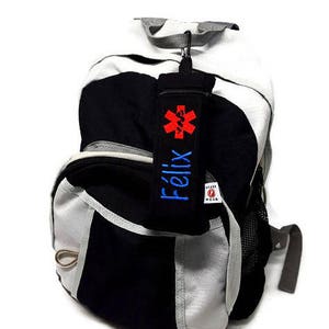 May include: Black and gray backpack with a red medical symbol and the name "Felix" embroidered on a blue strap.