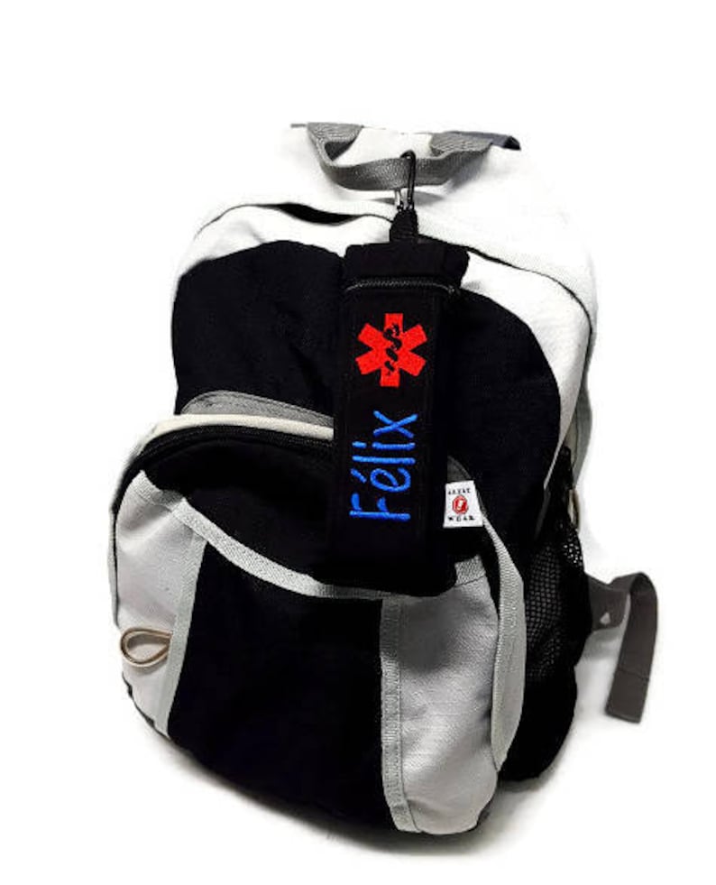 May include: Black and gray backpack with a red medical symbol and the name "Felix" embroidered on a blue strap.