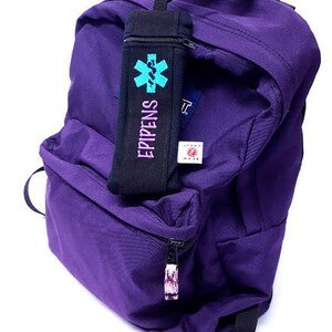 May include: Purple backpack with a black zippered pouch attached to the front. The pouch has a blue and green star of life symbol and the text "EPIPENS" printed on it.